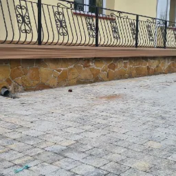 Wykończony taras z desek kompozytowych na podmurówce z kamienia naturalnego, z czarną, ozdobną balustradą. Widoczny fragment kostki brukowej i zaparkowane samochody w tle.