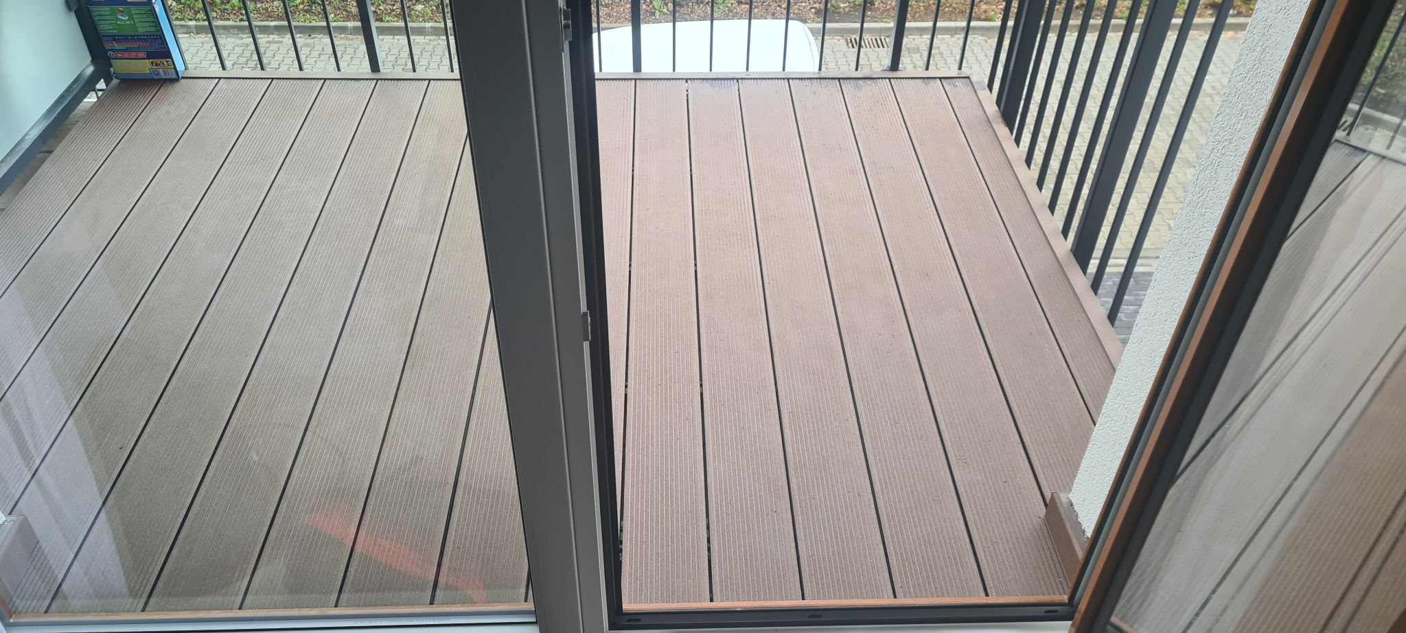 Balkon z brązową podłogą z desek kompozytowych, widok z wnętrza przez otwarte drzwi balkonowe, z widoczną barierką i fragmentem elewacji.