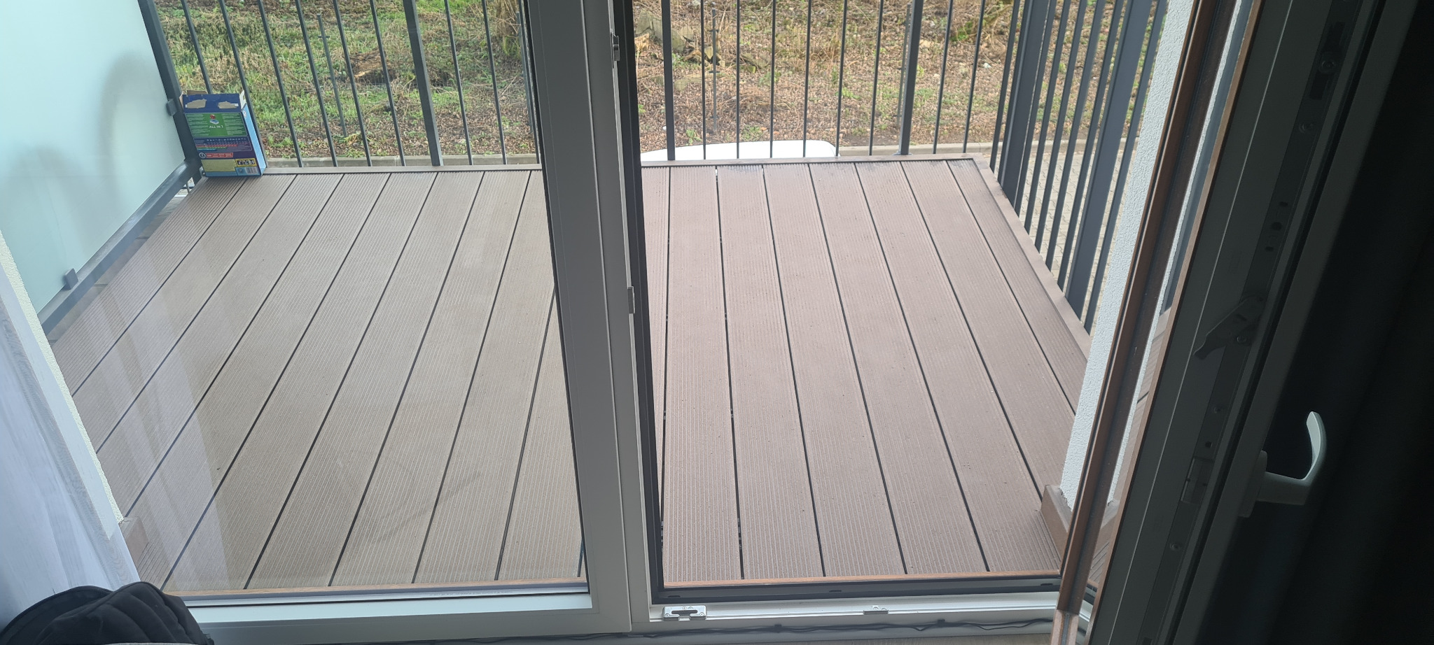 Balkon z brązową podłogą z desek kompozytowych, widok z wnętrza przez przesuwne drzwi balkonowe, na podłodze stoi karton.