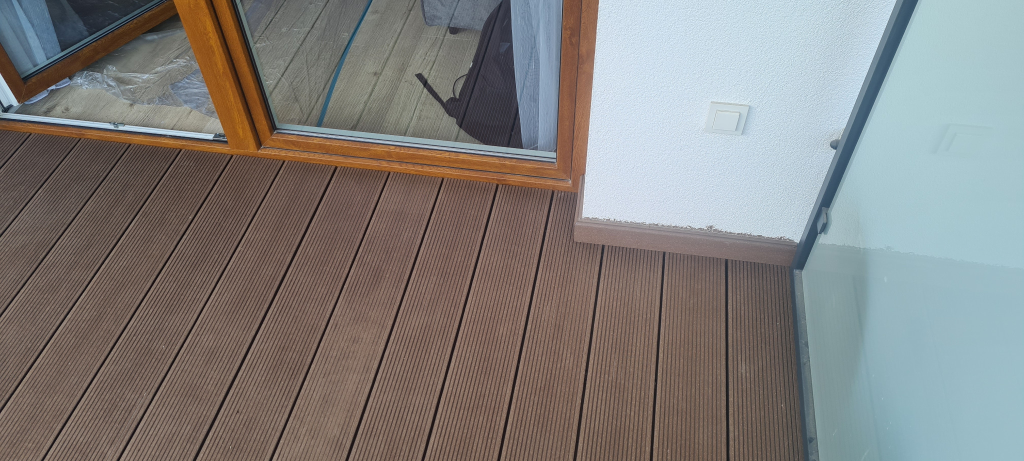 Nisko: Brązowa deska tarasowa kompozytowa ułożona przy oknie z brązową ramą i białą ścianą z gniazdkiem.