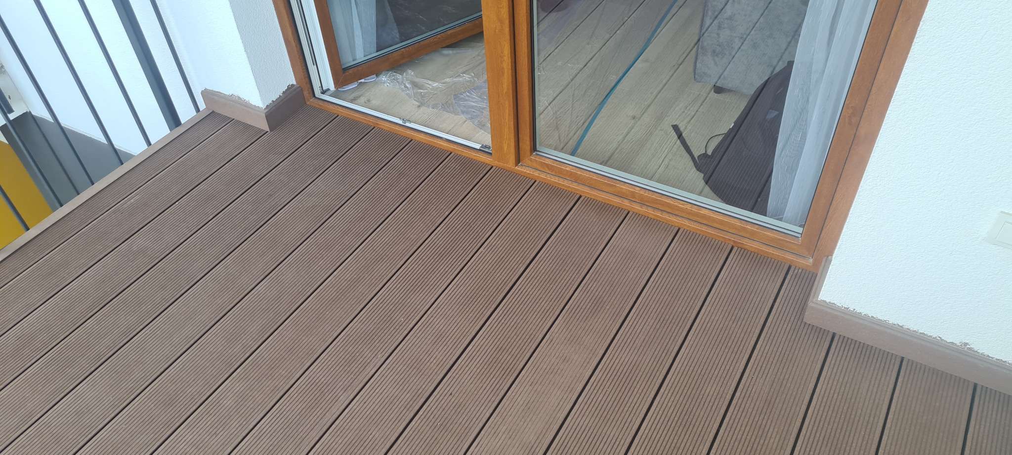 Nowoczesny balkon wyłożony brązowymi deskami z kompozytu, widok z góry na przesuwne okno balkonowe z drewnianą ramą.