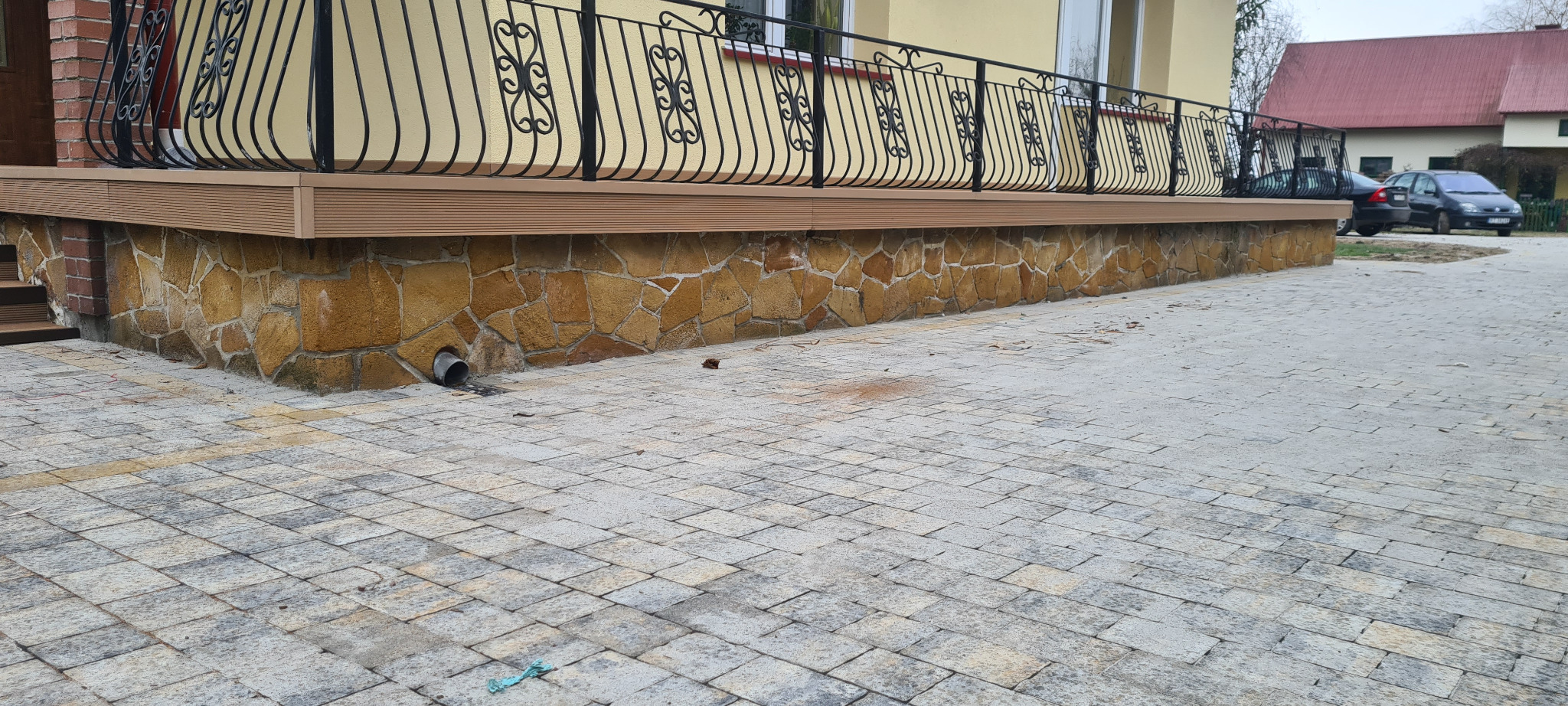 Wykończony taras z desek kompozytowych na podmurówce z kamienia naturalnego, z czarną, ozdobną balustradą. Widoczny fragment kostki brukowej i zaparkowane samochody w tle.