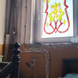 ARFO sp. z o.o. - Remont starego domu: widoczne kable elektryczne na ścianie z odpadającym tynkiem, okno ze zdobieniami kwiatowymi. Elementy instalacji i wystrój w trakcie modernizacji.
