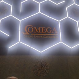 ARFO sp. z o.o. - Świecące heksagony tworzą logo 'OMEGA Ośrodek Szkolenia Zawodowego'. W tle ściana z zegarem i dwie osoby w recepcji.