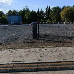 Szlaban parkingowy z automatycznym systemem kontroli dostępu na żwirowym parkingu, widoczne budynki w tle i spowalniacze na pierwszym planie.