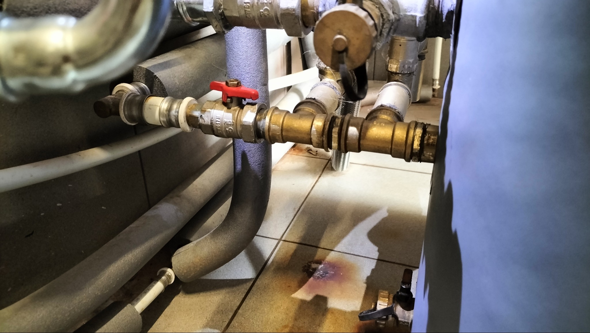 Złożony system rur miedzianych i plastikowych z zaworem z czerwoną rączką, widoczne ślady wycieku na kafelkach pod instalacją. Detal instalacji hydraulicznej.