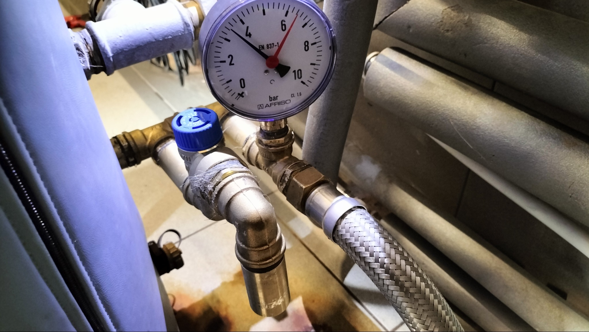 Manometr AFRISO na instalacji hydraulicznej z rurami miedzianymi i elastycznym przewodem w oplocie. Widoczne zawory z niebieskimi pokrętłami.