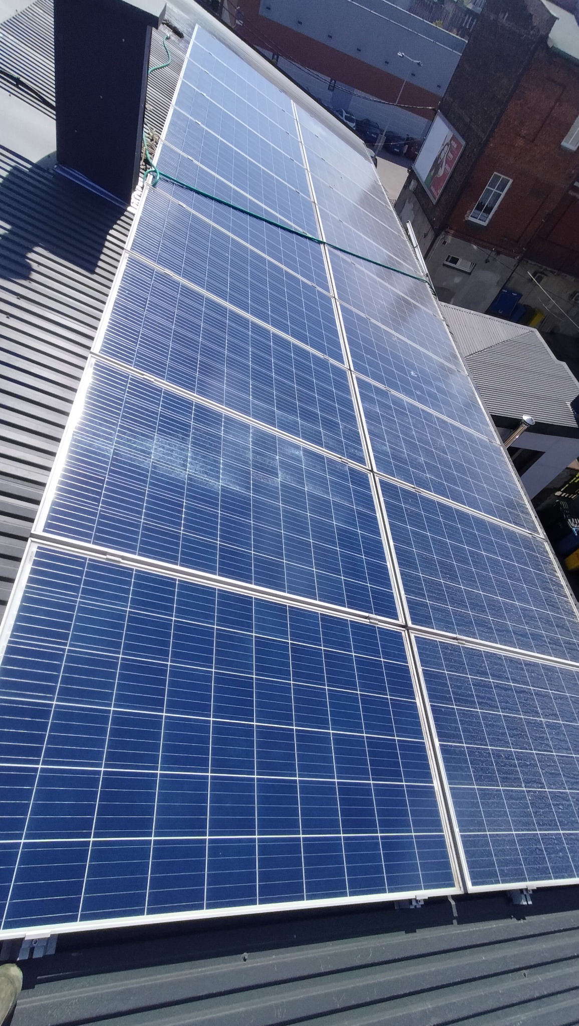 Instalacja paneli słonecznych na metalowym dachu, widok z góry. Panele w niebieskiej tonacji, z widocznymi liniami podziału. W tle budynki miejskie.