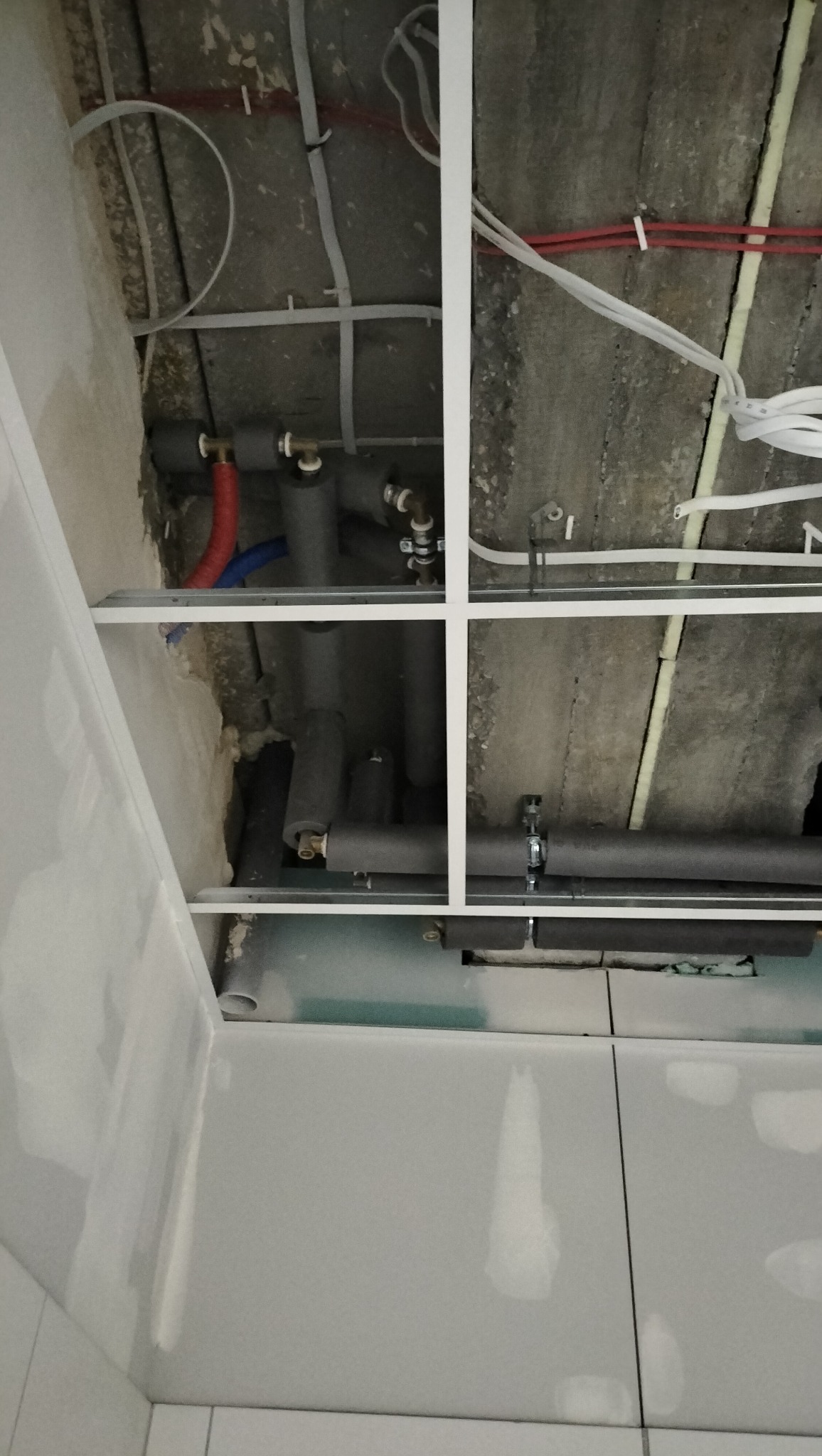 Szare rury instalacji sanitarnej z izolacją termiczną pod sufitem podwieszanym, widoczne przewody elektryczne i fragment ściany z płyt gipsowych.