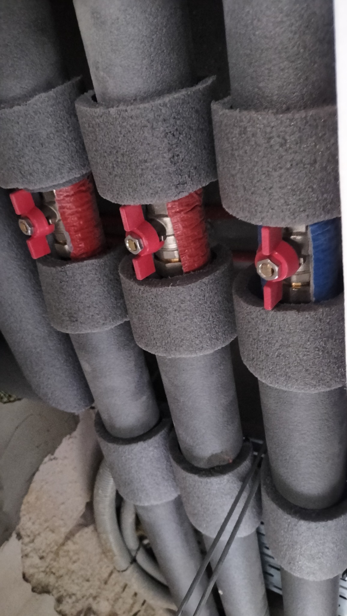 Szare rury grzewcze z izolacją i czerwonymi zaworami, ułożone równolegle, widoczne od dołu pod kątem, z fragmentem ściany w tle.