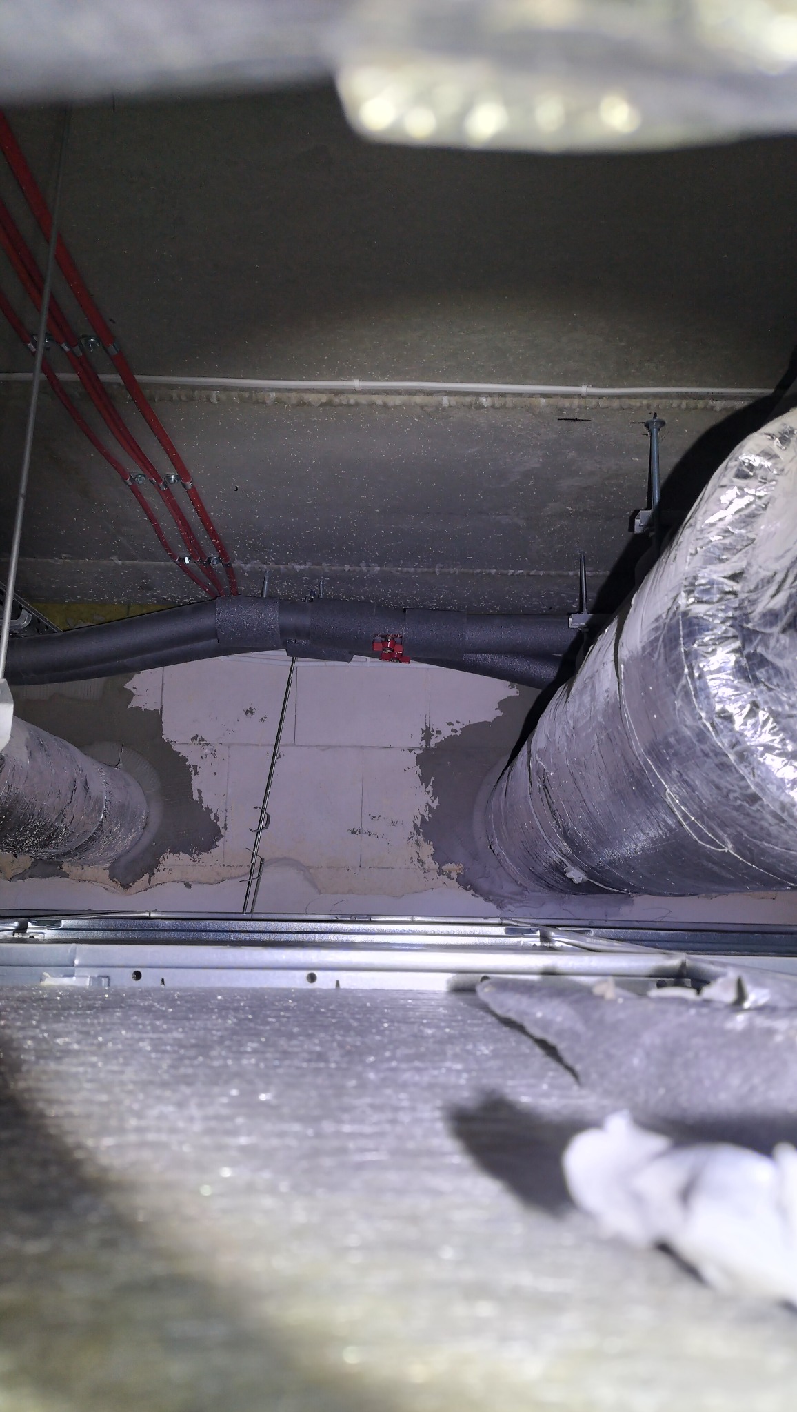 Ukończona instalacja wentylacyjna pod sufitem z izolowanymi rurami i czerwonymi przewodami, widok z dołu, w trakcie montażu sufitu podwieszanego.