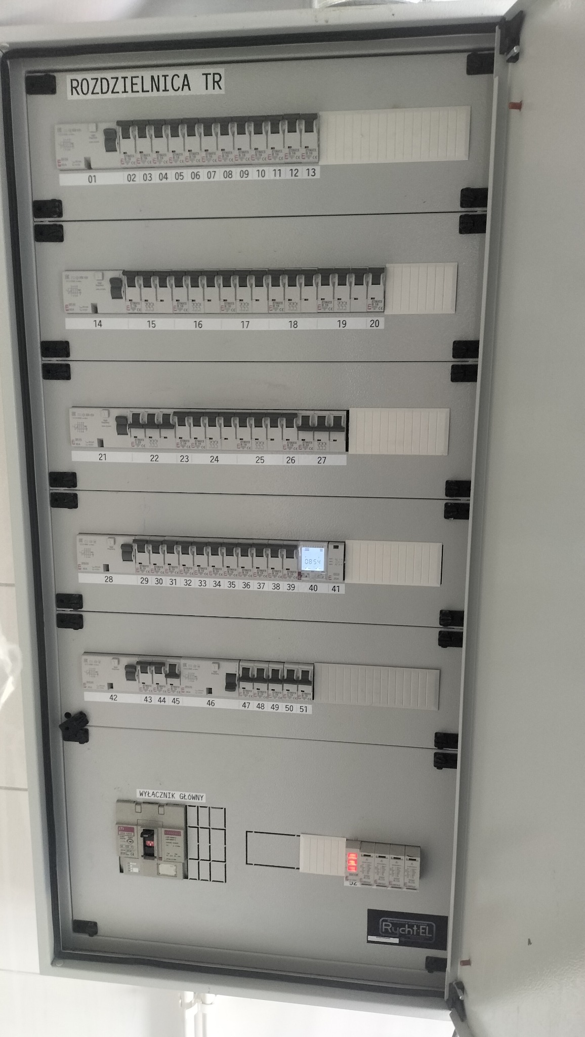 Otwarta skrzynka elektryczna, rozdzielnica TR z numerowanymi wyłącznikami, widoczny wyłącznik główny i zabezpieczenia. Szara obudowa, białe oznaczenia.
