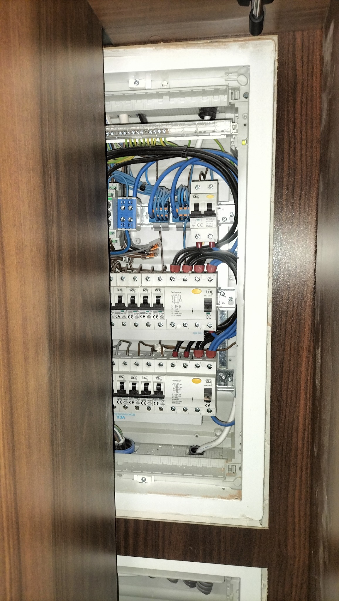 Otwarta skrzynka elektryczna z bezpiecznikami, widoczne kable w kolorach niebieskim i czarnym. Elementy instalacji zamontowane w szafce w kolorze drewna.
