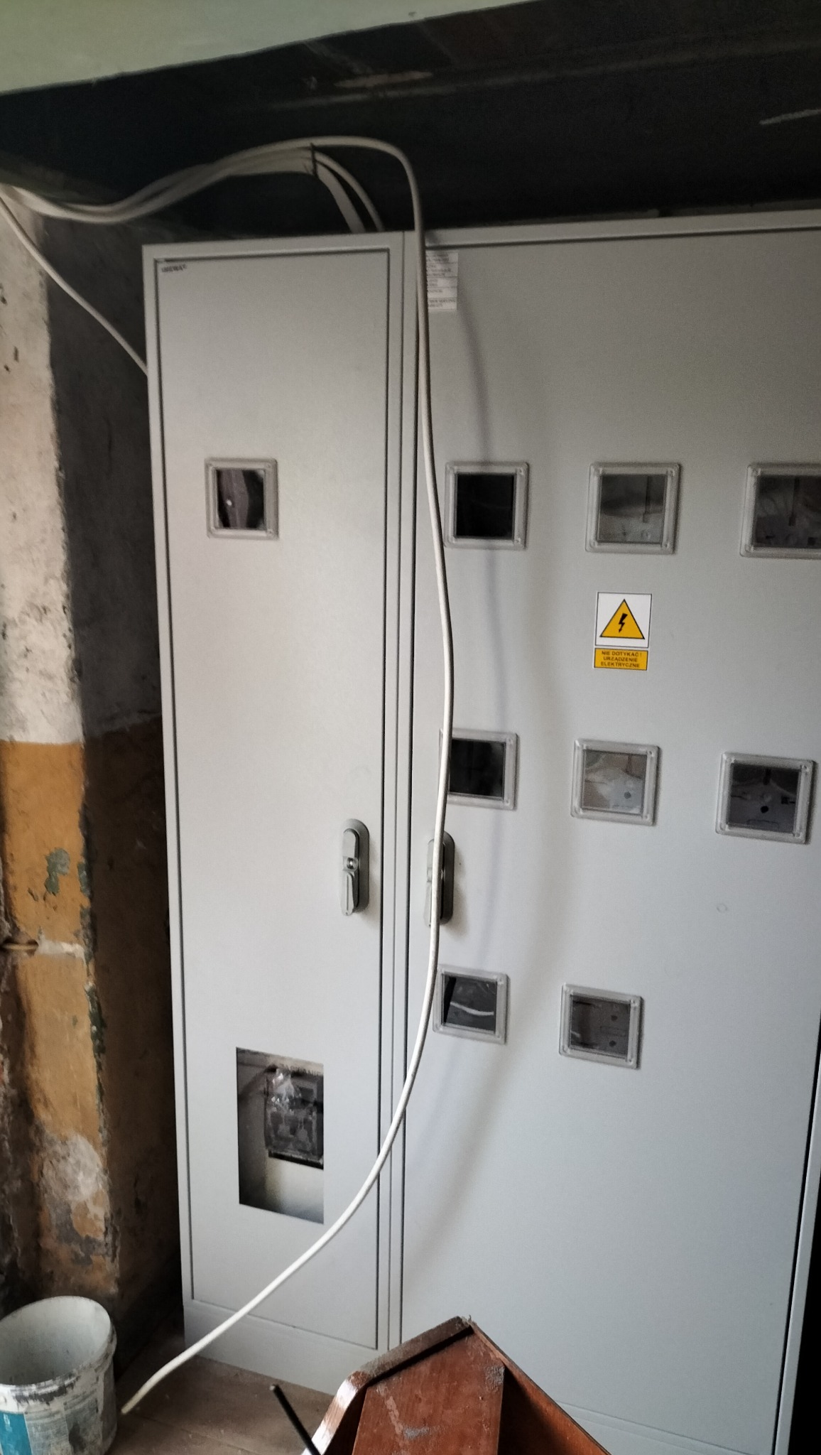 Szara szafa elektryczna z okienkami i ostrzeżeniem. Widoczne kable, fragment ściany z odpadającym tynkiem i wiadro. Ujęcie z dołu, wnętrze w trakcie prac.