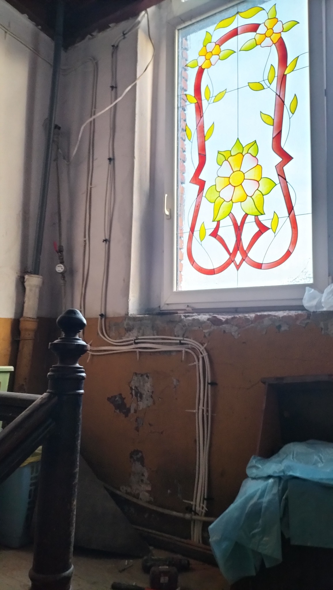 Remont starego domu: widoczne kable elektryczne na ścianie z odpadającym tynkiem, okno ze zdobieniami kwiatowymi. Elementy instalacji i wystrój w trakcie modernizacji.
