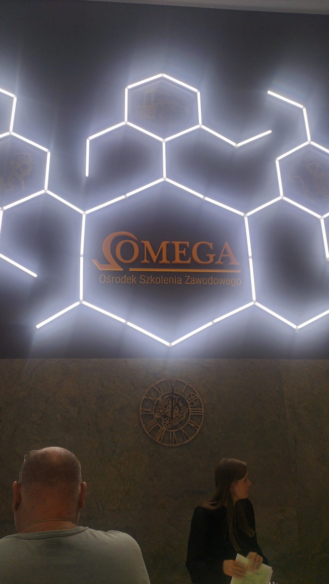 Świecące heksagony tworzą logo 'OMEGA Ośrodek Szkolenia Zawodowego'. W tle ściana z zegarem i dwie osoby w recepcji.