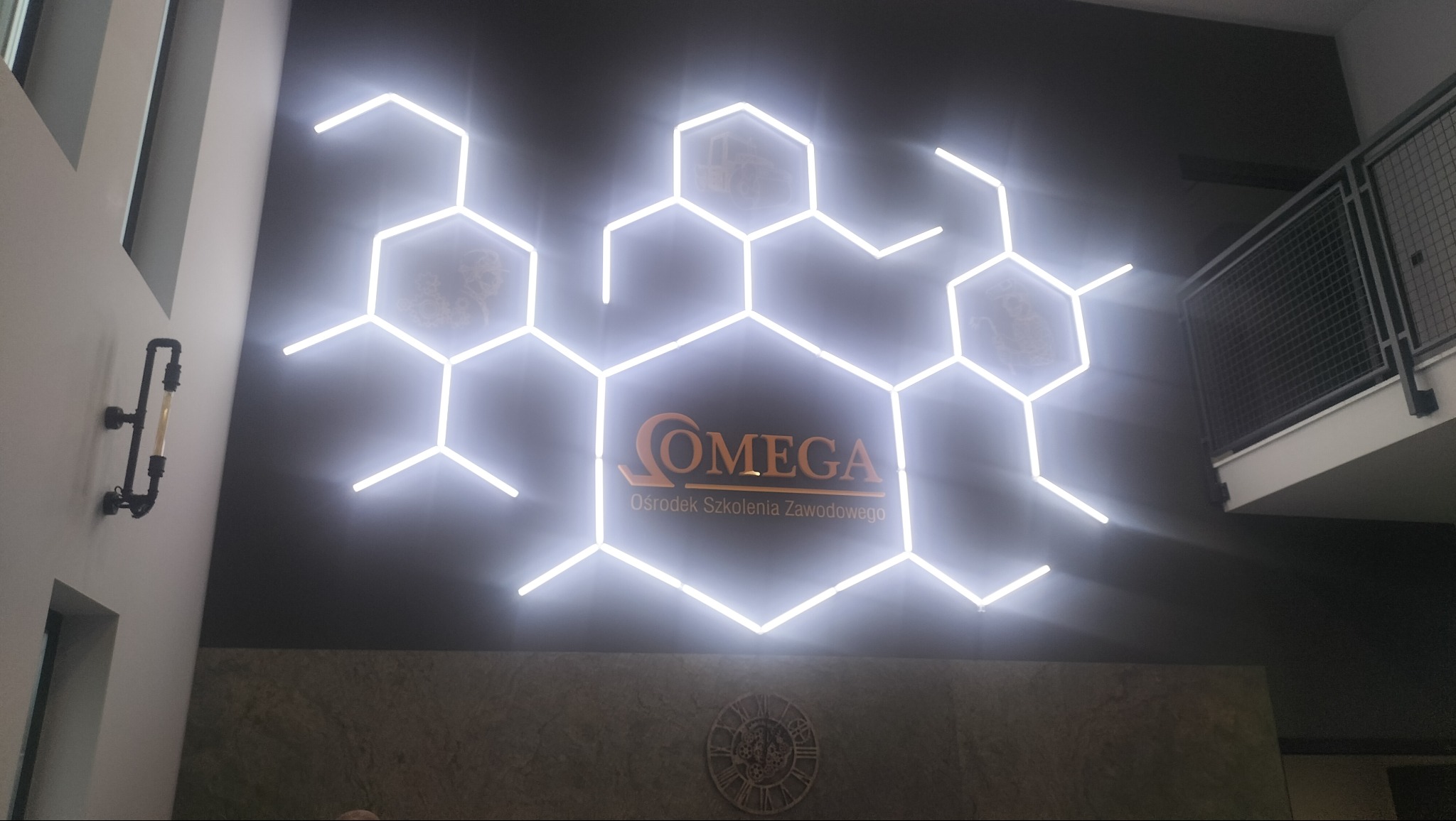 Oświetlenie LED w kształcie heksagonów, tworzące logo 'OMEGA Ośrodek Szkolenia Zawodowego' na ciemnej ścianie. Dodatkowa lampa w stylu industrialnym po lewej.