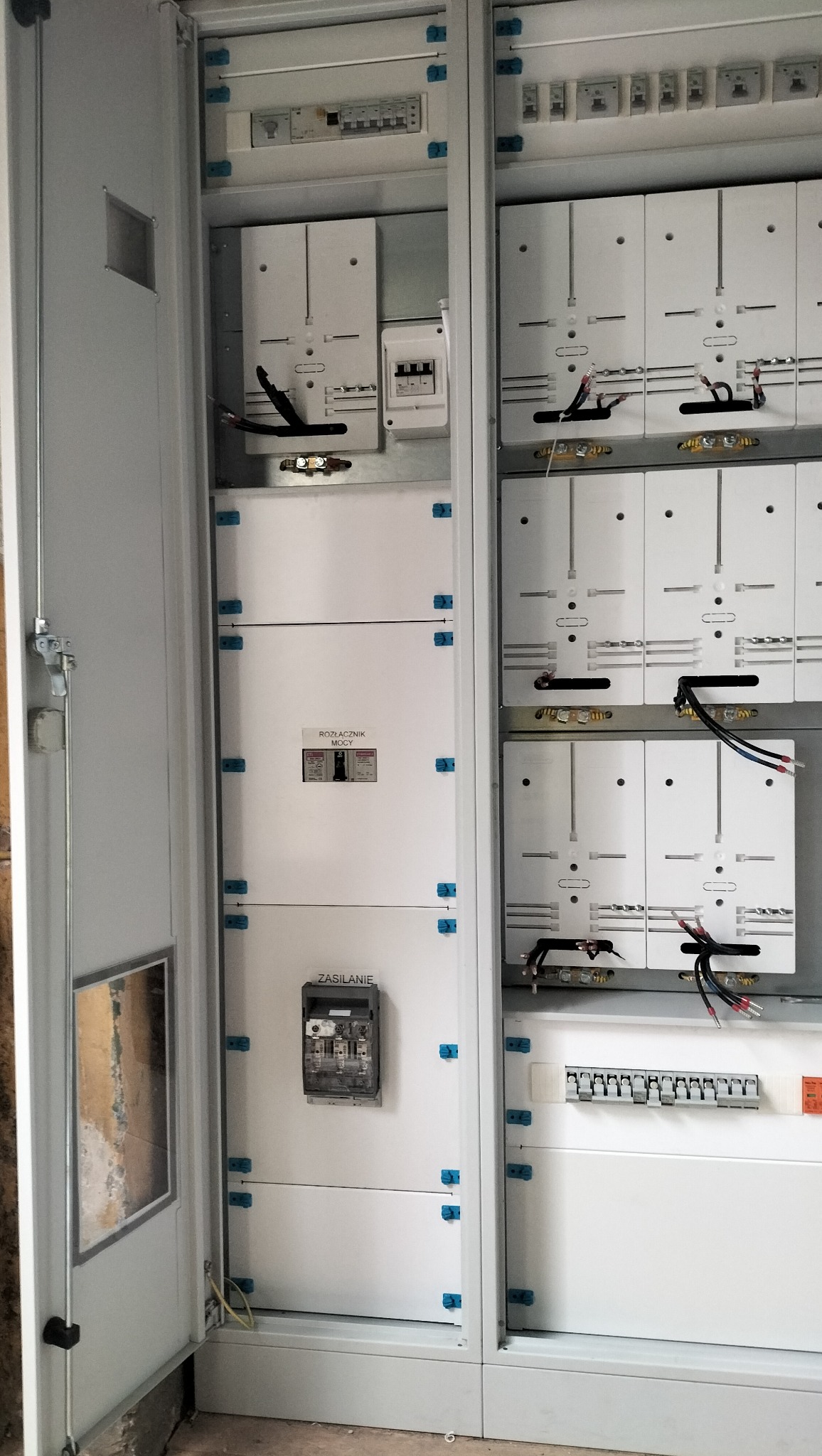 Rozdzielnica elektryczna z otwartymi drzwiami, widoczne zabezpieczenia, wyłączniki i miernik energii. Biała obudowa z niebieskimi elementami mocującymi.