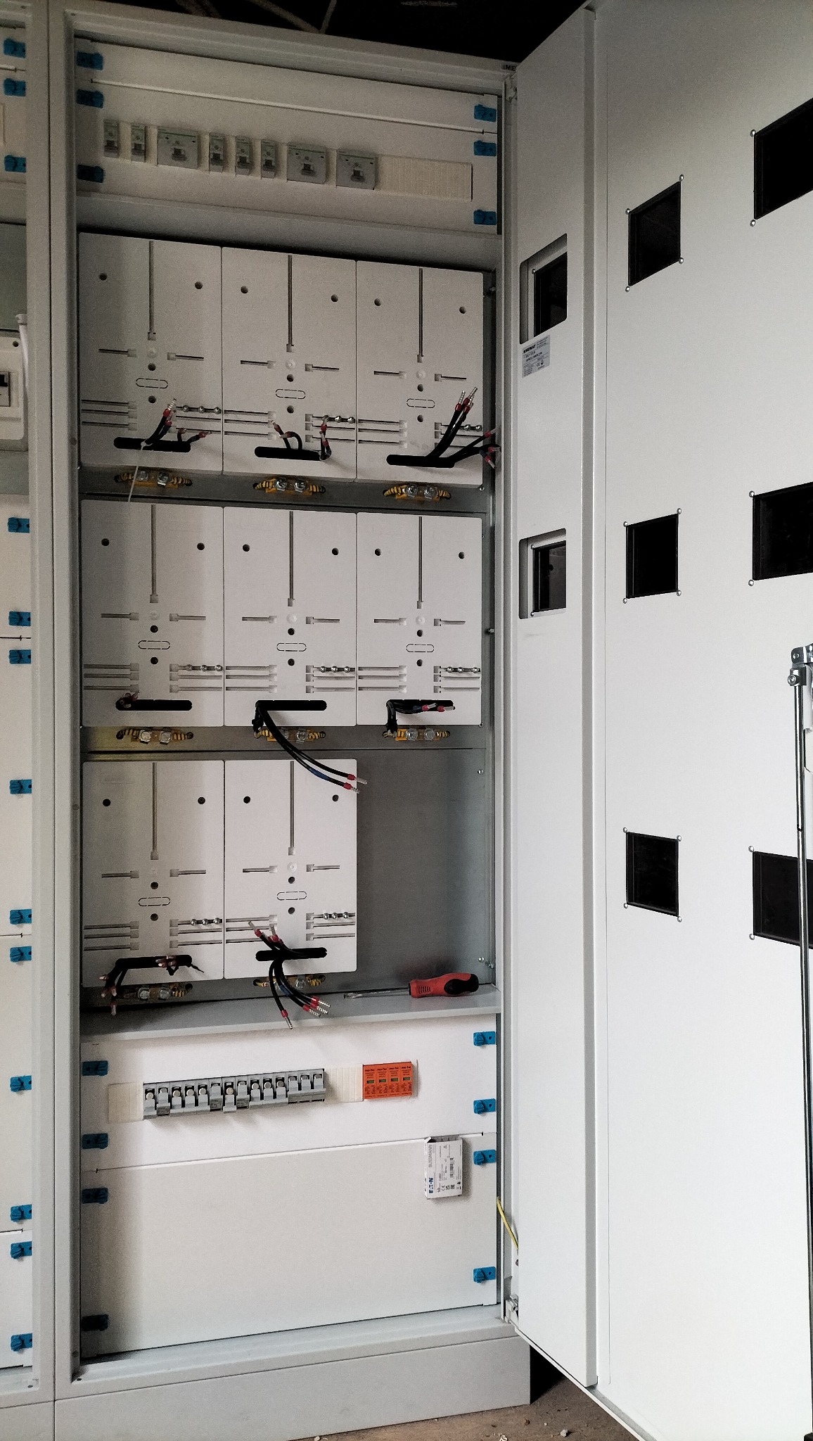 Otwarta szafa elektryczna z widocznymi bezpiecznikami, przewodami i elementami instalacji. Widoczne narzędzie - śrubokręt. Biała obudowa z kwadratowymi otworami.