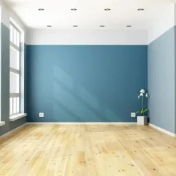 Puste, jasne pomieszczenie z drewnianą podłogą, ścianą w odcieniach błękitu i szarości oraz białymi oknami. Minimalistyczny wystrój z pojedynczym kwiatem w doniczce.