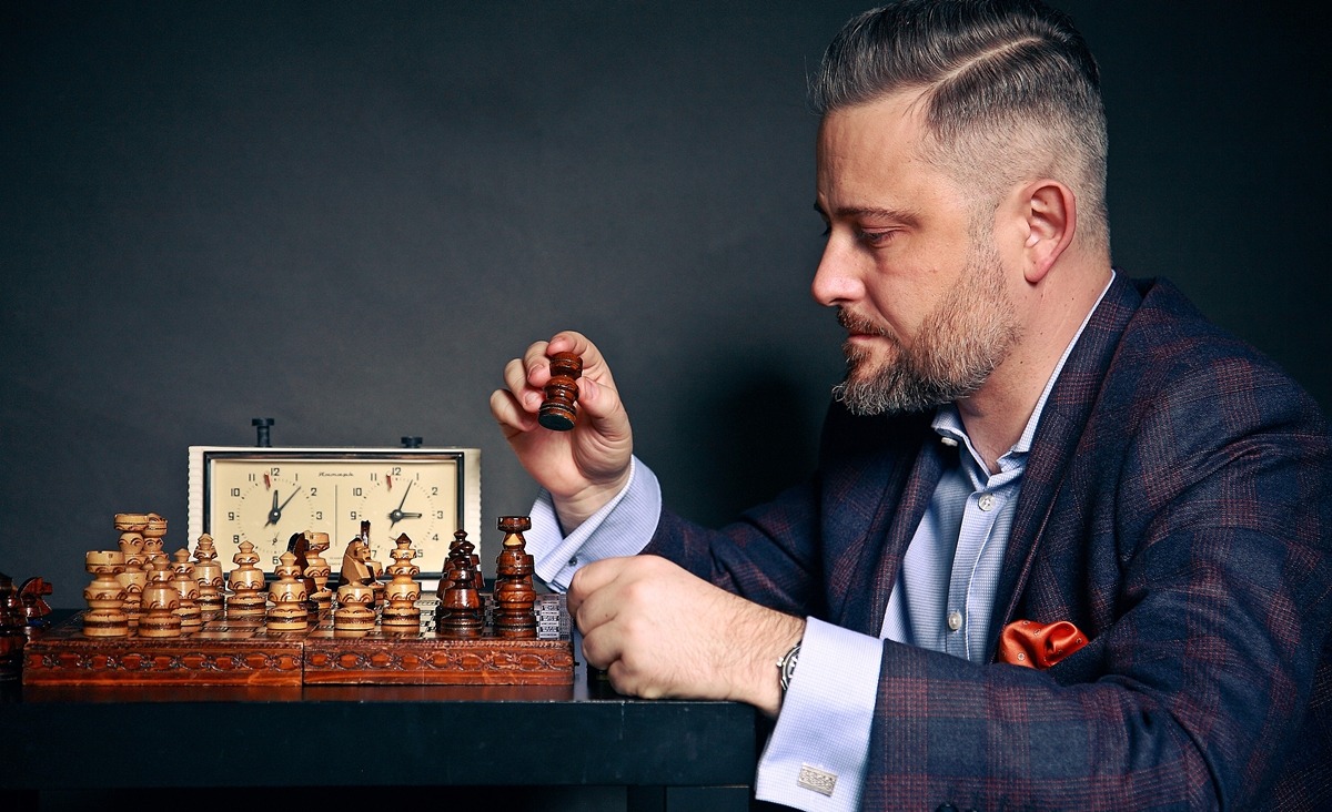 Mężczyzna w garniturze, zamyślony, trzyma pionka szachowego nad szachownicą z drewnianymi figurami, w tle zegar szachowy.