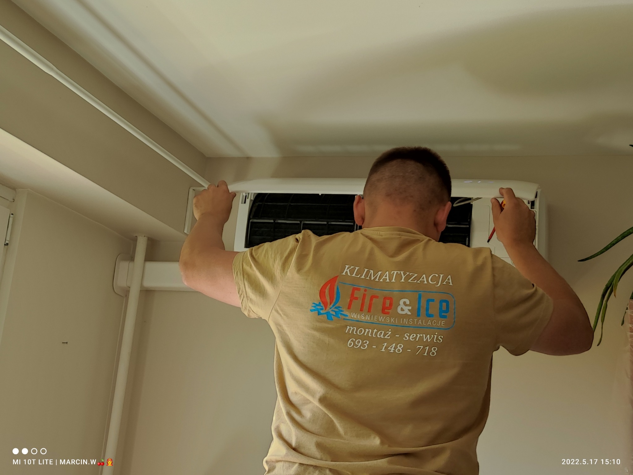 Instalator w koszulce z logo firmy Fire&Ice serwisuje klimatyzator na ścianie, używając śrubokręta.