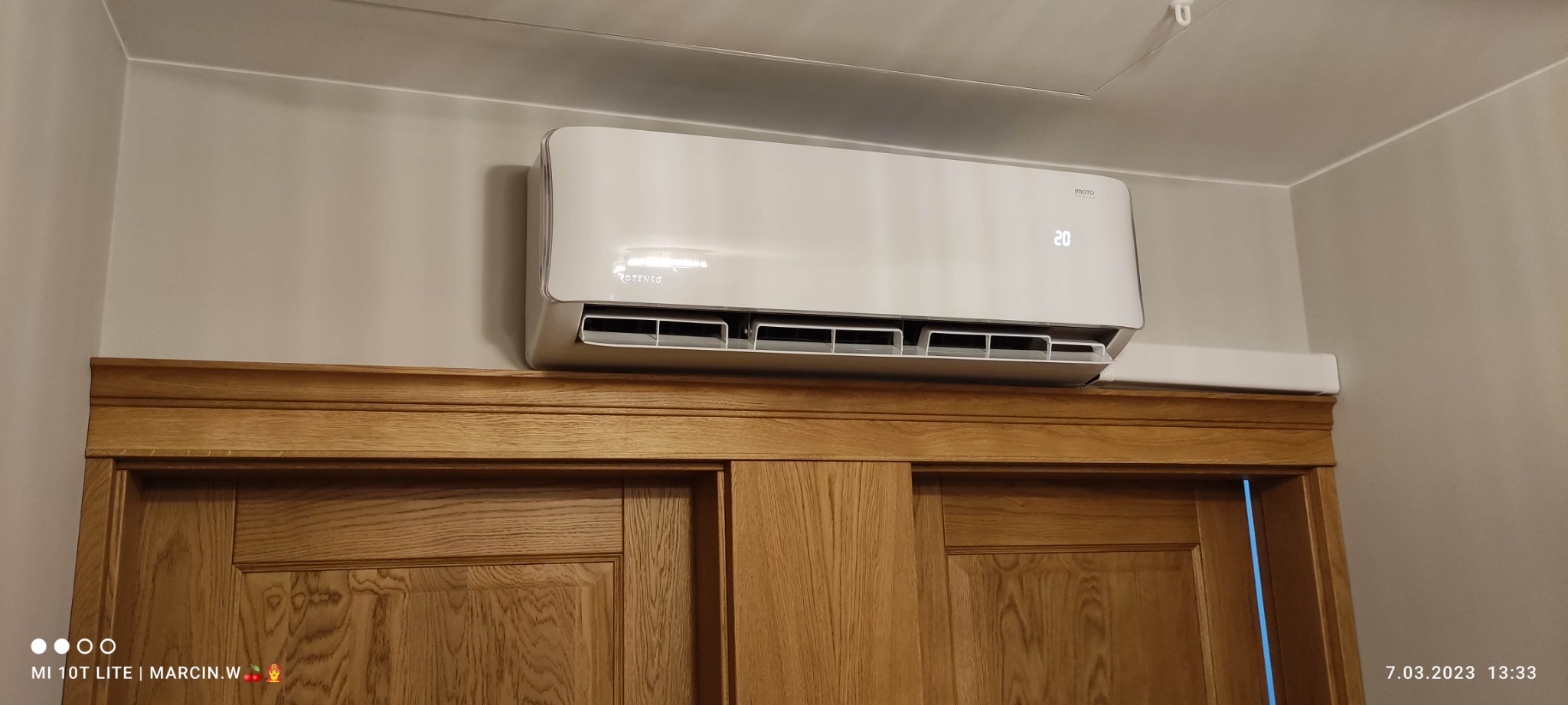 Biały klimatyzator Rotenso zamontowany nad drewnianymi drzwiami przesuwnymi w pomieszczeniu.