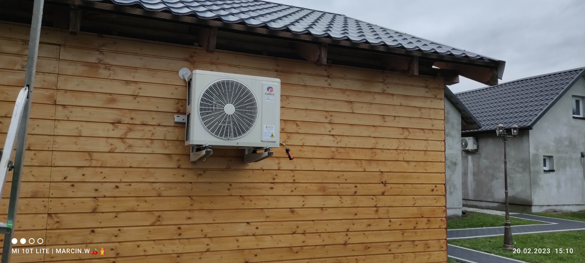 Biała jednostka zewnętrzna klimatyzatora marki Gree zamontowana na drewnianej ścianie budynku.