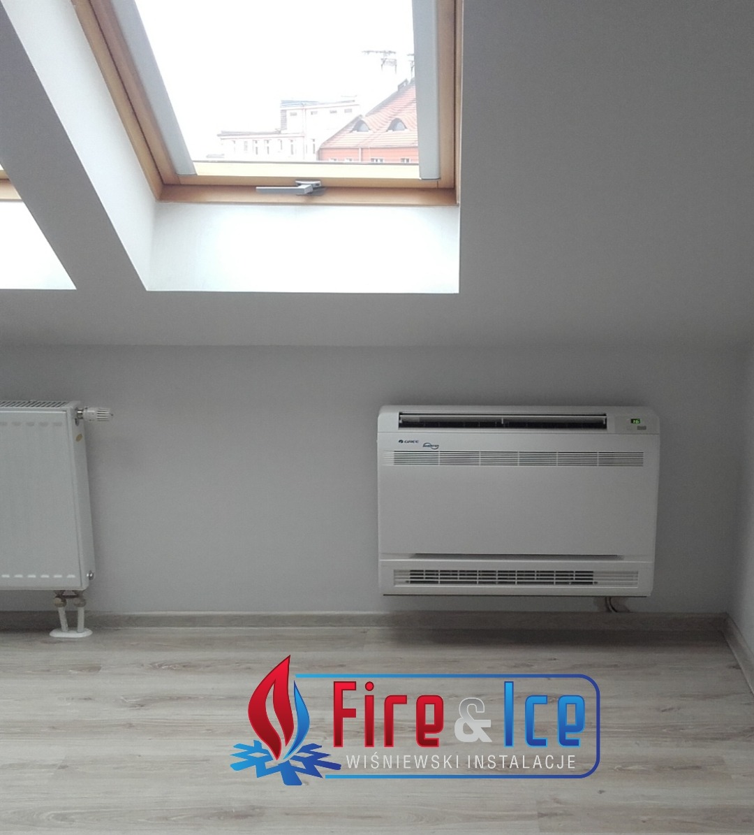 Biała, przypodłogowa jednostka klimatyzacyjna Gree, zainstalowana w szarym pokoju na poddaszu z oknami dachowymi, obok grzejnika.