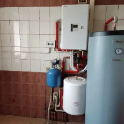 Instalacja kotła gazowego Toshiba z trzema zbiornikami: niebieskim, białym i szarym Vasti, z widocznymi rurami miedzianymi i plastikowymi w pomieszczeniu z brązowo-białymi płytkami.