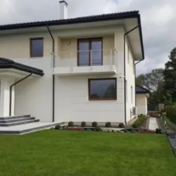 Dwukondygnacyjny dom z beżowo-białą elewacją, brązowymi ramami okiennymi i szarą kostką na schodach wejściowych, otoczony świeżo przystrzyżonym trawnikiem i zadbanym ogrodem.