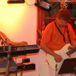 Kobieta śpiewa do mikrofonu grając na keyboardzie Roland E-50, obok mężczyzna gra na białej gitarze elektrycznej podczas występu na scenie.