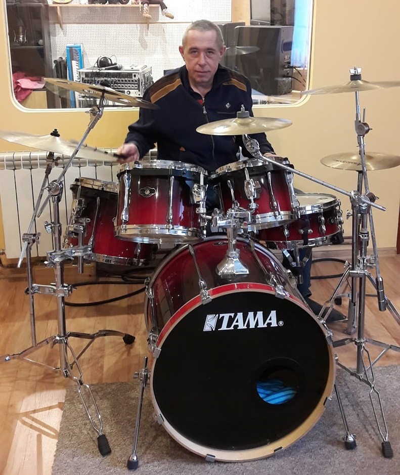 Perkusista pozuje za zestawem perkusyjnym Tama w pomieszczeniu z radiatorem i sprzętem audio w tle.