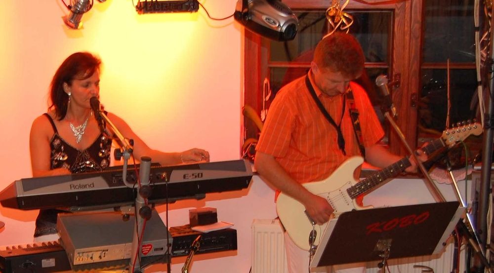 Kobieta śpiewa do mikrofonu grając na keyboardzie Roland E-50, obok mężczyzna gra na białej gitarze elektrycznej podczas występu na scenie.