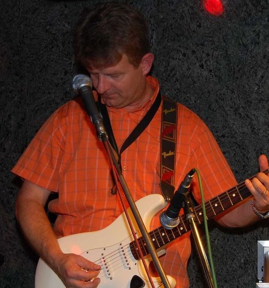 Mężczyzna w pomarańczowej koszuli gra na białej gitarze elektrycznej, śpiewając do mikrofonu na tle ciemnej ściany.