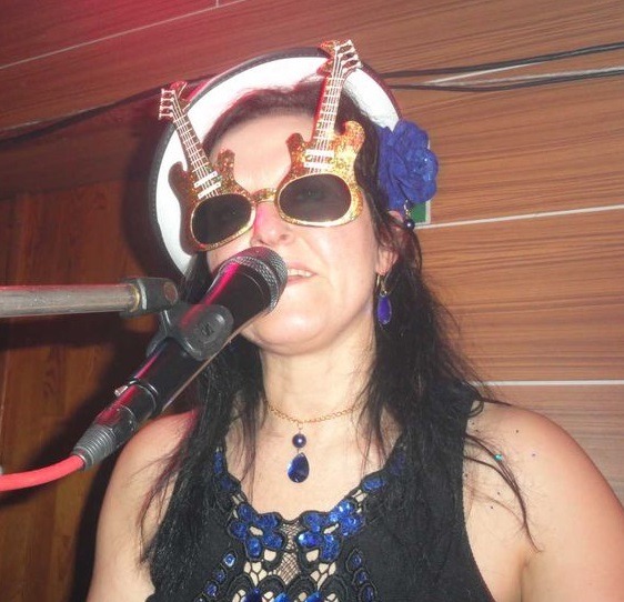 Wokalistka z mikrofonem, w okularach w kształcie złotych gitar, z niebieskim kwiatem we włosach, śpiewa na scenie.