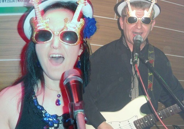 Wokalistka z niebieskim kwiatem we włosach i gitarzysta śpiewają do mikrofonów, oboje w okularach w kształcie gitary, mężczyzna gra na białej gitarze elektrycznej.