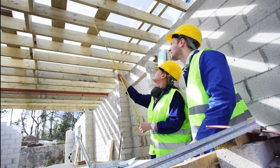 Dwoje pracowników budowlanych w żółtych kaskach i odblaskowych kamizelkach mierzy odległość między drewnianymi belkami stropu za pomocą zwijanej miarki, na pierwszym planie laptop i metalowa listwa...