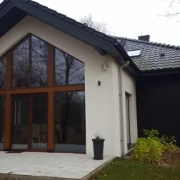 Nowoczesny dom z dużymi oknami i drzwiami przesuwnymi, brązowe ramy, jasna elewacja i ciemny dach, widok z zewnątrz na taras wyłożony jasnymi płytkami.