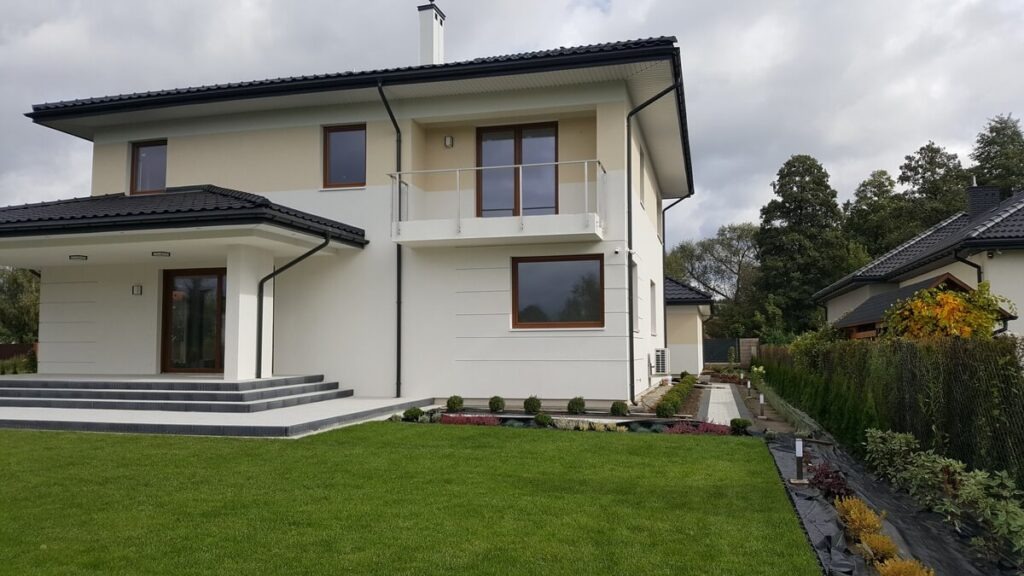 Nowoczesny dom jednorodzinny z brązowymi oknami, balkonem ze szklaną balustradą i zadbanym trawnikiem.