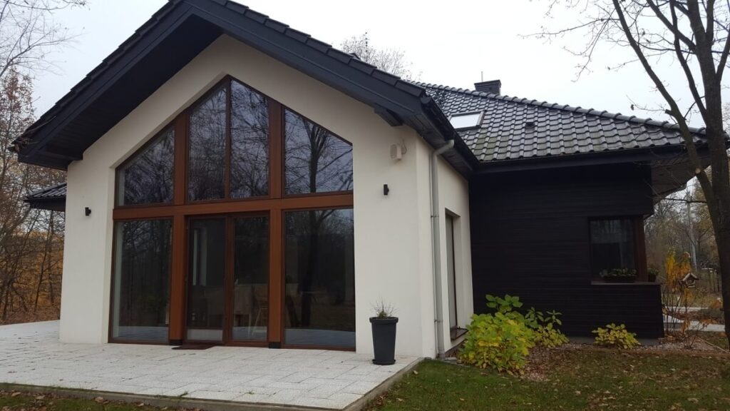 Nowoczesny dom z dużymi oknami i drzwiami przesuwnymi, brązowe ramy, jasna elewacja i ciemny dach, widok z zewnątrz na taras wyłożony jasnymi płytkami.