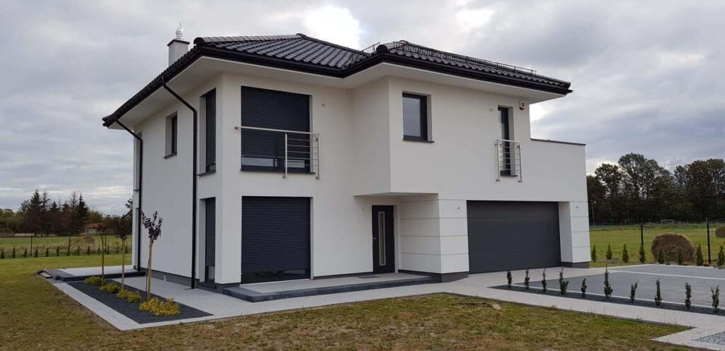 Nowoczesny, dwukondygnacyjny dom z białymi ścianami, czarnym dachem i grafitowymi roletami zewnętrznymi oraz bramą garażową, otoczony zadbanym trawnikiem i dekoracyjnymi nasadzeniami.