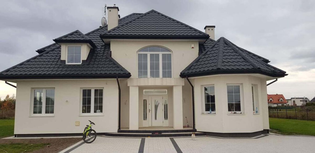 Elewacja domu jednorodzinnego z czarną dachówką, białymi oknami PCV i drzwiami wejściowymi, z rowerem i małym psem na podjeździe.