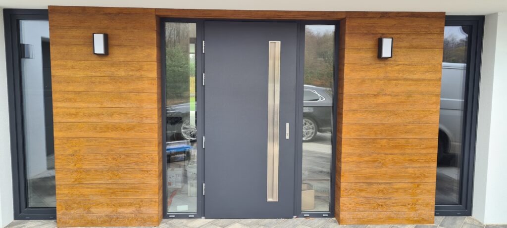 Nowoczesne drzwi wejściowe w kolorze antracytowym z podłużnym, metalowym uchwytem, osadzone w drewnianej elewacji z oknami po bokach, oświetlone dwoma kinkietami.