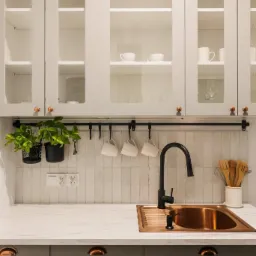 Nowoczesna kuchnia w odcieniach bieli i szarości z miedzianym zlewem i czarną armaturą, wiszące kubki i zioła w doniczkach na relingu, przeszklone szafki górne z porcelaną.