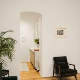 Jasne wnętrze mieszkania z drewnianą podłogą, zielonymi roślinami, ciemnozielonymi fotelami i obrazkiem na ścianie, widok na kuchnię przez łukowe przejście.