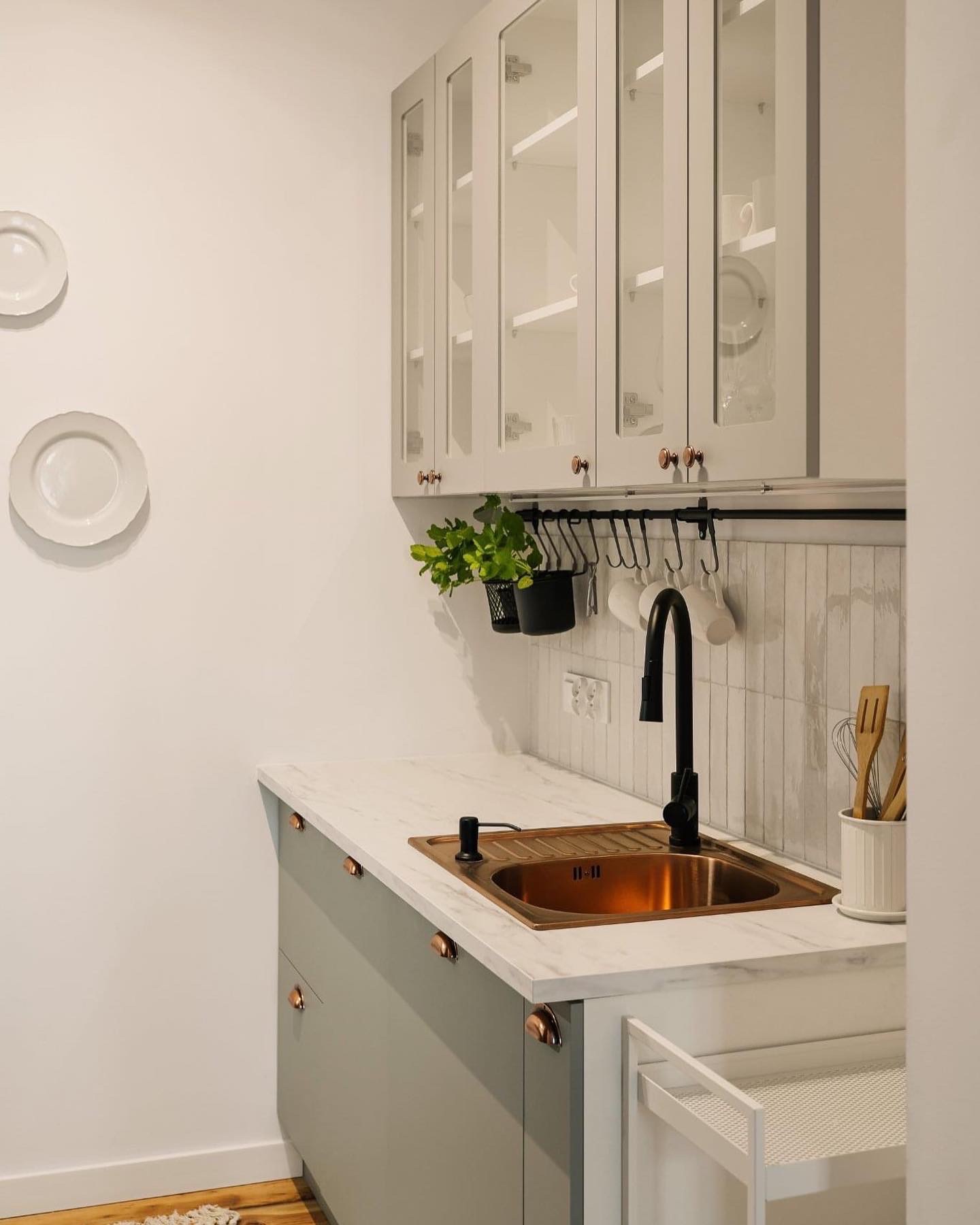 Nowoczesna kuchnia z szarymi szafkami, miedzianym zlewem i czarną armaturą, uzupełniona o wiszące kubki i zioła w czarnych doniczkach.
