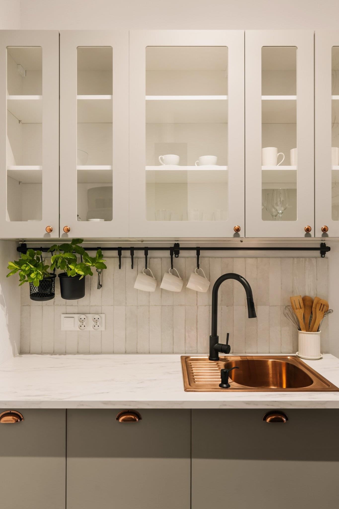 Nowoczesna kuchnia w odcieniach bieli i szarości z miedzianym zlewem i czarną armaturą, wiszące kubki i zioła w doniczkach na relingu, przeszklone szafki górne z porcelaną.