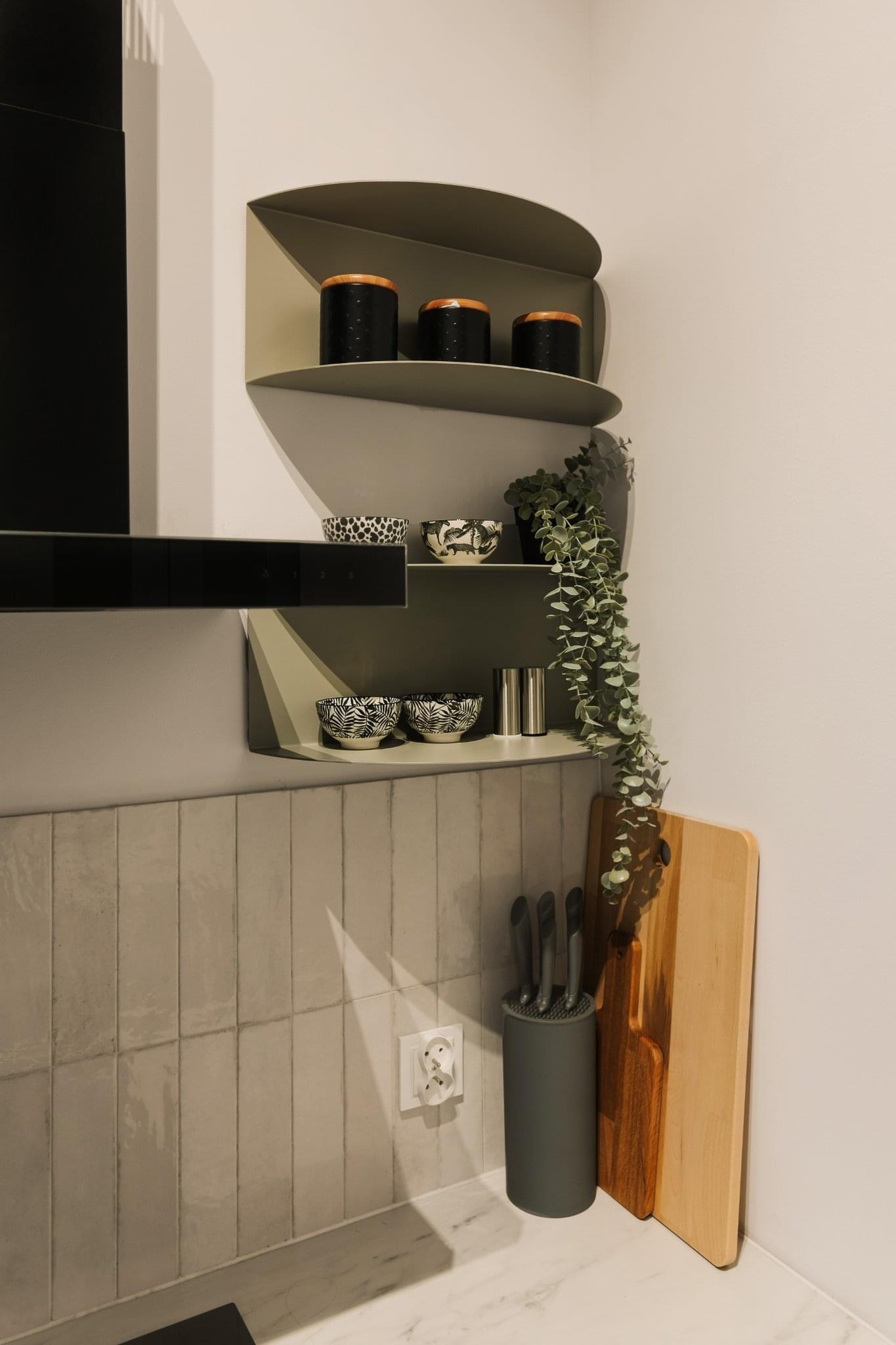 Kącik kuchenny z półkami w odcieniach szarości, na których stoją czarne pojemniki z drewnianymi pokrywkami, miseczki w czarno-białe wzory, srebrne pojemniki na przyprawy i zwisająca roślina.