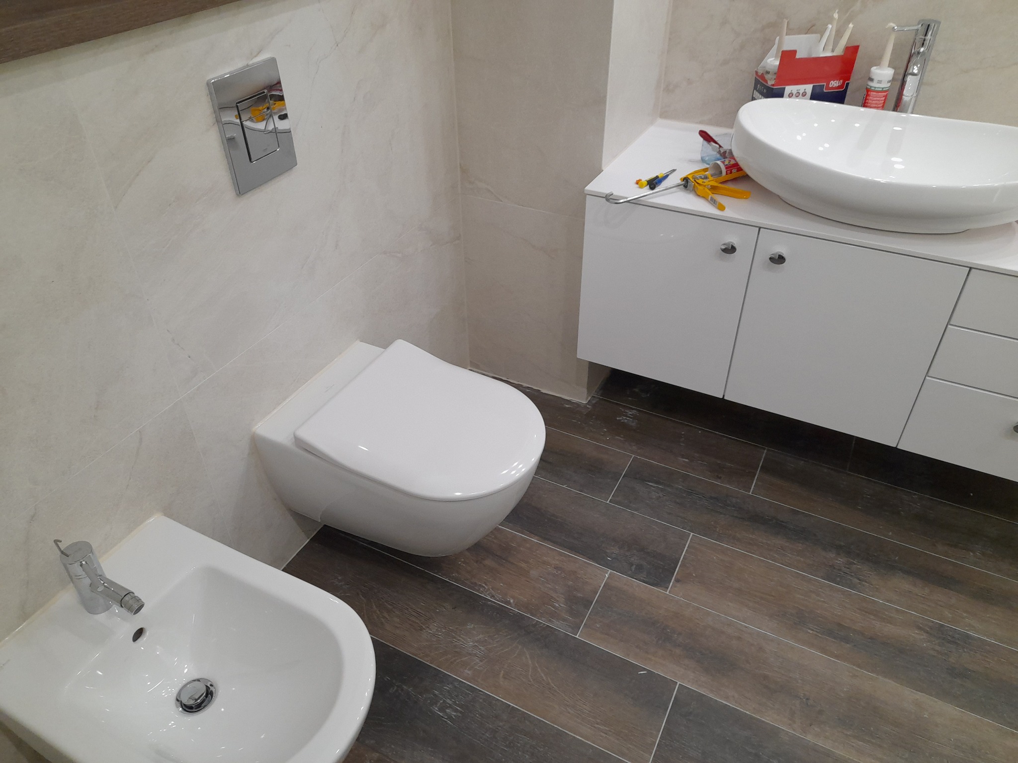 Wykończona łazienka z podwieszaną miską WC, bidetem, umywalką nablatową i szafką pod umywalką, płytki imitujące drewno na podłodze, akcesoria montażowe na blacie.